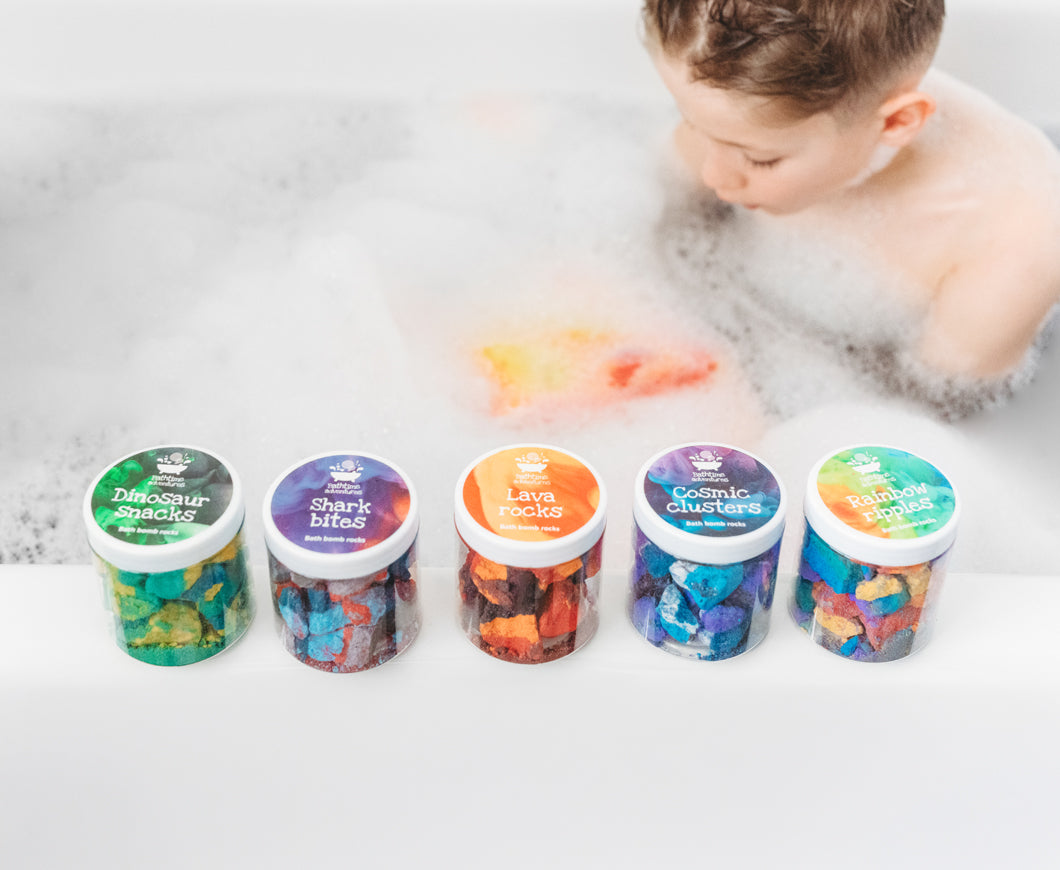 Five colourful pots of bath bomb rocks in front of child sitting in bubbly bath with colourful water