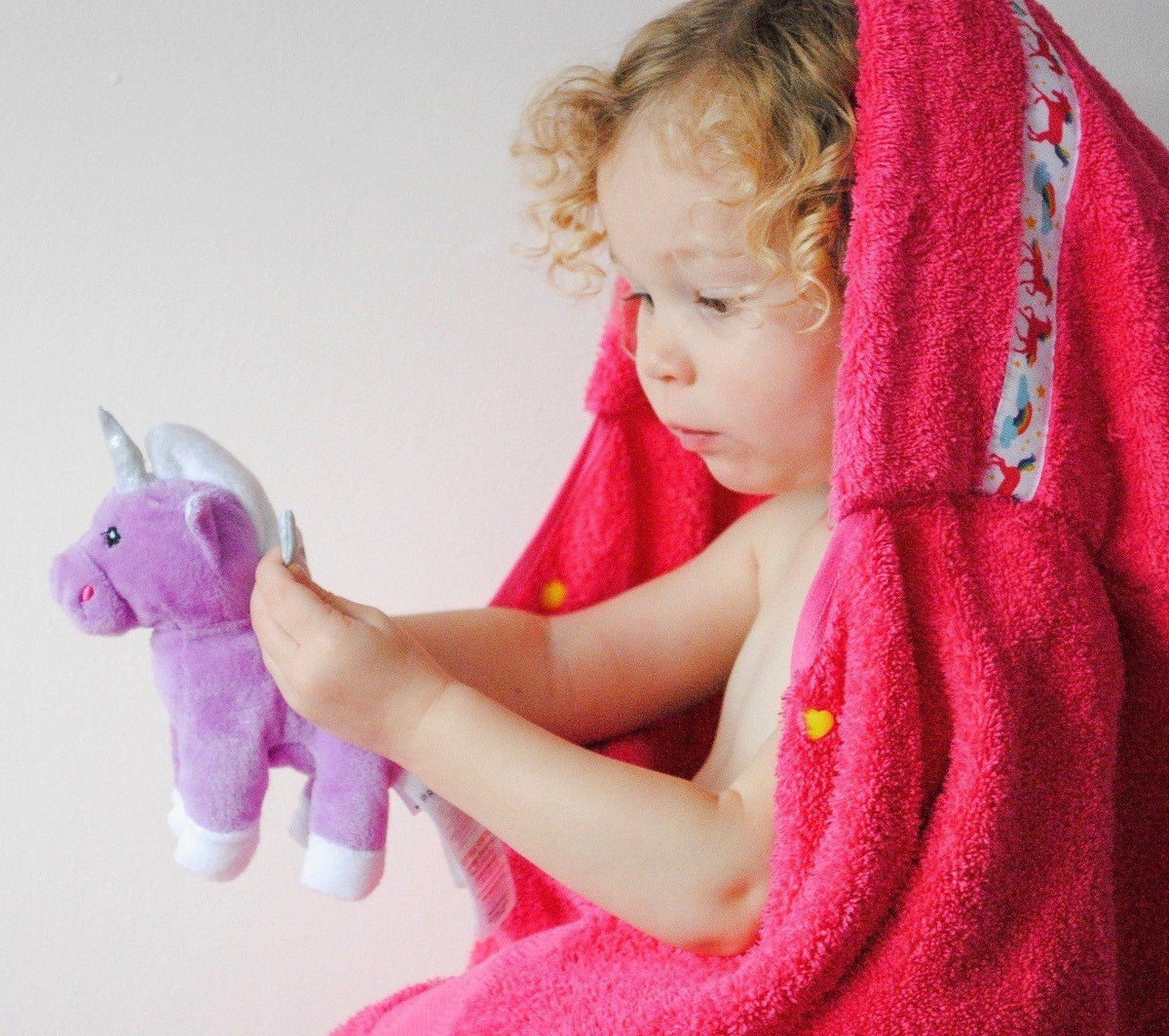 Child wearing pink hooded towel with Unicorns trim whilst holding a unicorn soft toy
